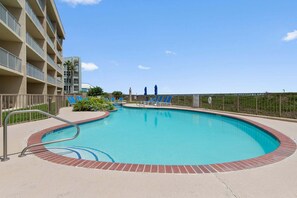 A swimming pool with a brick edge, surrounded by a fence, and a building with balconies in the background.