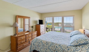 A bedroom with a bed, dresser, mirror, and a view of a pool through the window.