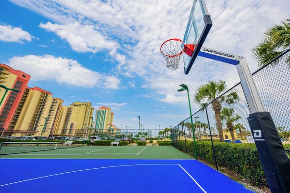 Ein Basketballplatz im Freien mit einem Netz mit der Aufschrift DOMINATOR, umgeben von einem Maschendrahtzaun, Palmen und Gebäuden im Hintergrund.
