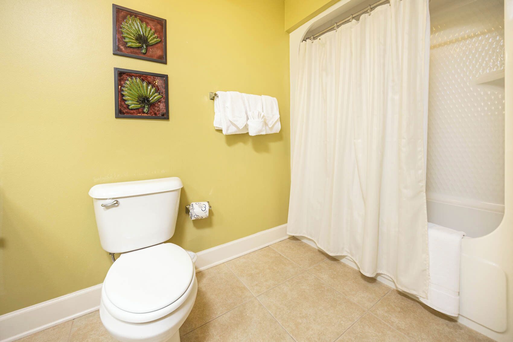 A bathroom with a toilet, two framed pictures on the wall, a white towel on a rack, and a white shower curtain.