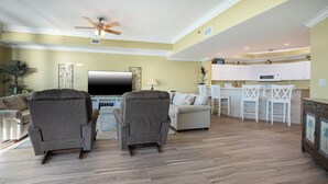 Una sala de estar con dos sillones, un sofá, un televisor de pantalla plana, un ventilador de techo y una zona de cocina con armarios blancos y taburetes de bar.