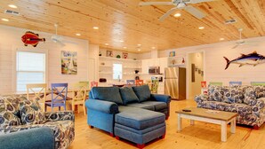 A spacious living room with a large blue sectional sofa, a matching armchair, and a wooden coffee table. The room features wooden floors, a ceiling fan, and a kitchen area visible through an open door. - You are certainly Shore To Love It! This fabulous island style home is located r (Port Aransas)
