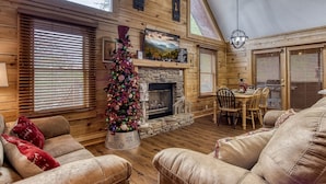 A cozy cabin interior with a fireplace, wooden walls, a dining area, and a Christmas tree. - Sleepy Bear | 2 King Suites • Hot Tub • Soaking Tub • Near Parkway • Eagles Ridge Resort (Pigeon Forge)