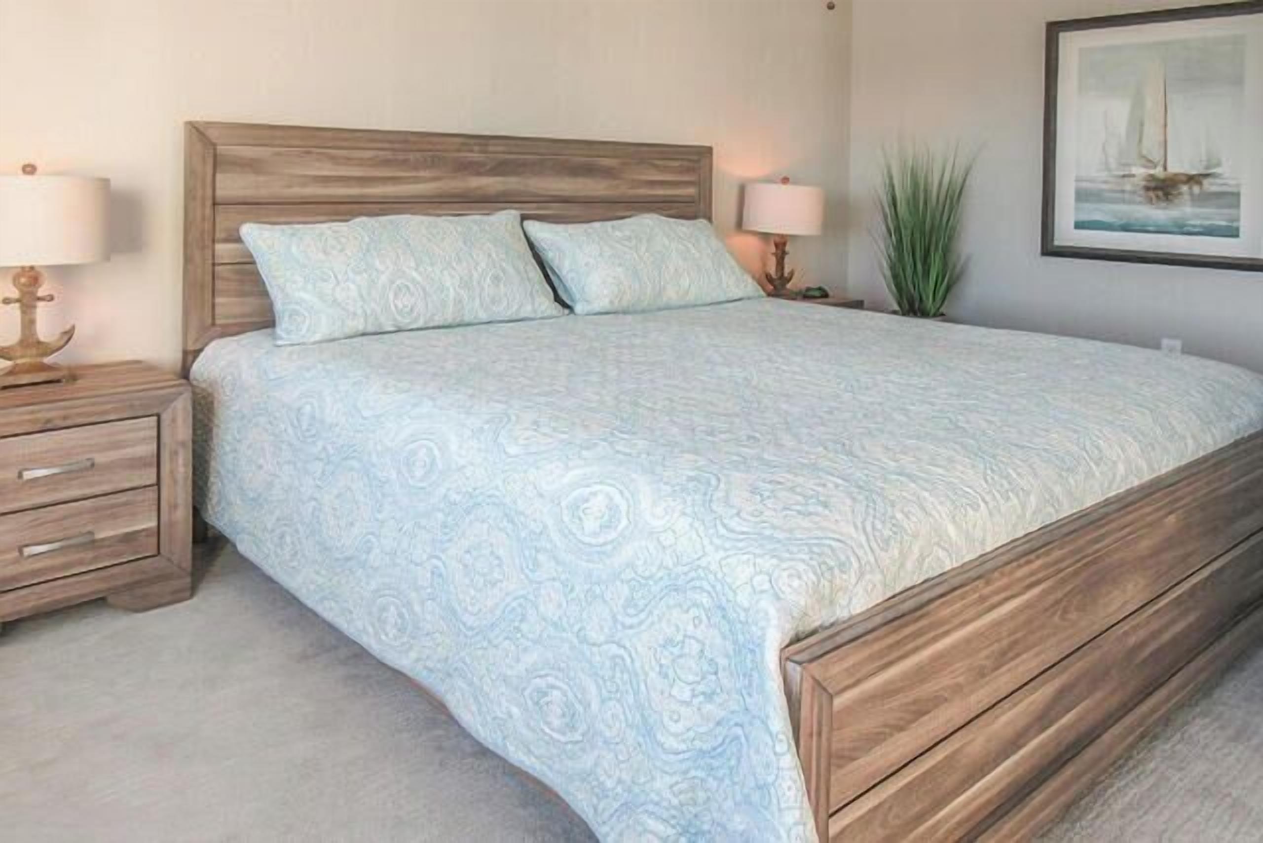 Un dormitorio con cabecera de madera, colcha estampada, dos mesitas de noche con lámparas y un cuadro enmarcado en la pared.