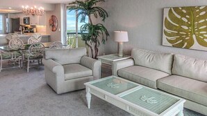 Un salon comprenant un canapé, deux fauteuils, une table basse en verre, une lampe, une plante et un tableau accroché au mur.