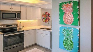 Une cuisine avec des meubles blancs, un four à micro-ondes, un four et un évier. Deux tableaux représentant des ananas sont accrochés au mur.