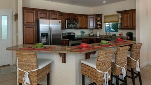 A kitchen with wooden cabinets, a stainless steel refrigerator, a stove, and a sink. There are four wicker bar stools with white cushions.