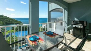 A balcony with a glass table set for two, a grill, and a view of the sea and greenery.