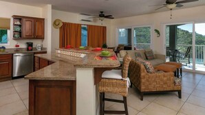A spacious kitchen with a central island, a dining area with wicker furniture, and a balcony with a view of the hills.