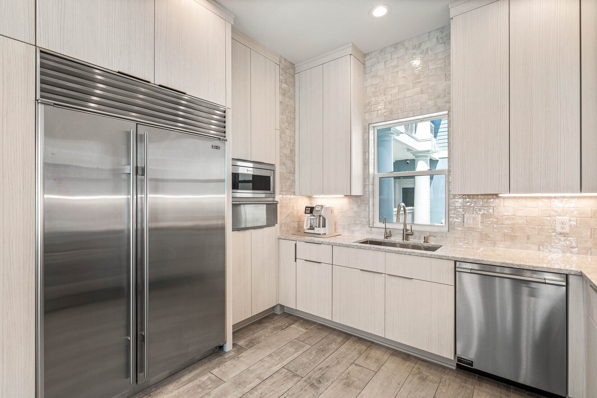 A modern kitchen with stainless steel appliances, light wood cabinetry, and a window overlooking a building.