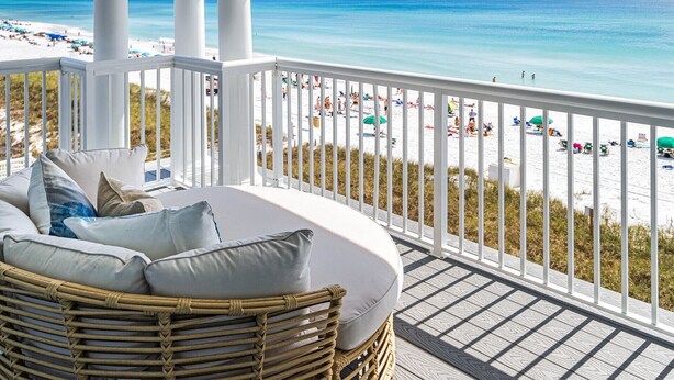 Ein Balkon mit einem Korbstuhl und Kissen, der Blick auf einen Strand mit Personen und Sonnenschirmen freigibt.