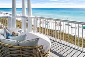 A balcony with a wicker chair and cushions, overlooking a beach with people and umbrellas. - SALER SHORES: 4-Story Gulf Front Home w/ Pool, Game Room, Elevator in Private Community! (Miramar Beach)