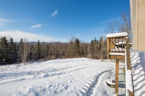 Una terraza de madera con una silla cubierta de nieve, un sendero nevado y un bosque al fondo.