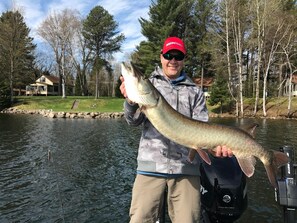 A person holding a large fish by a lake with a house in the background.