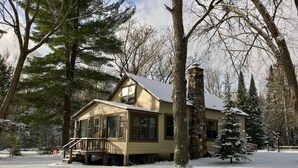 Une maison à deux étages avec un porche, entourée d’un paysage enneigé et d’arbres.