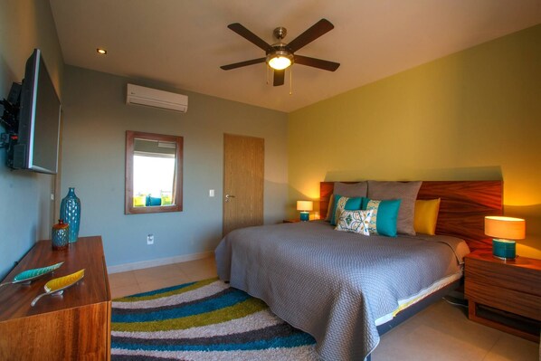 A bedroom with a bed, a ceiling fan, a TV, a mirror, a wooden dresser, and a colorful rug.