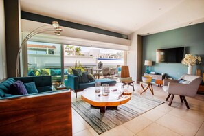 A modern living room with a sofa, coffee table, and chairs. There is a large window with a view of buildings and plants outside.
