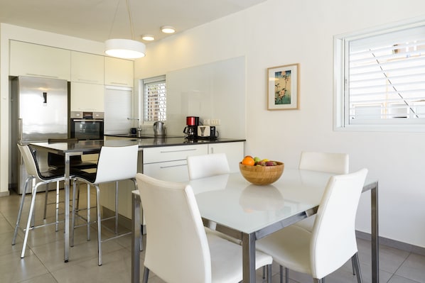 A modern kitchen with a dining area, featuring a stainless steel refrigerator, a coffee maker, and a bowl of fruit on the table.