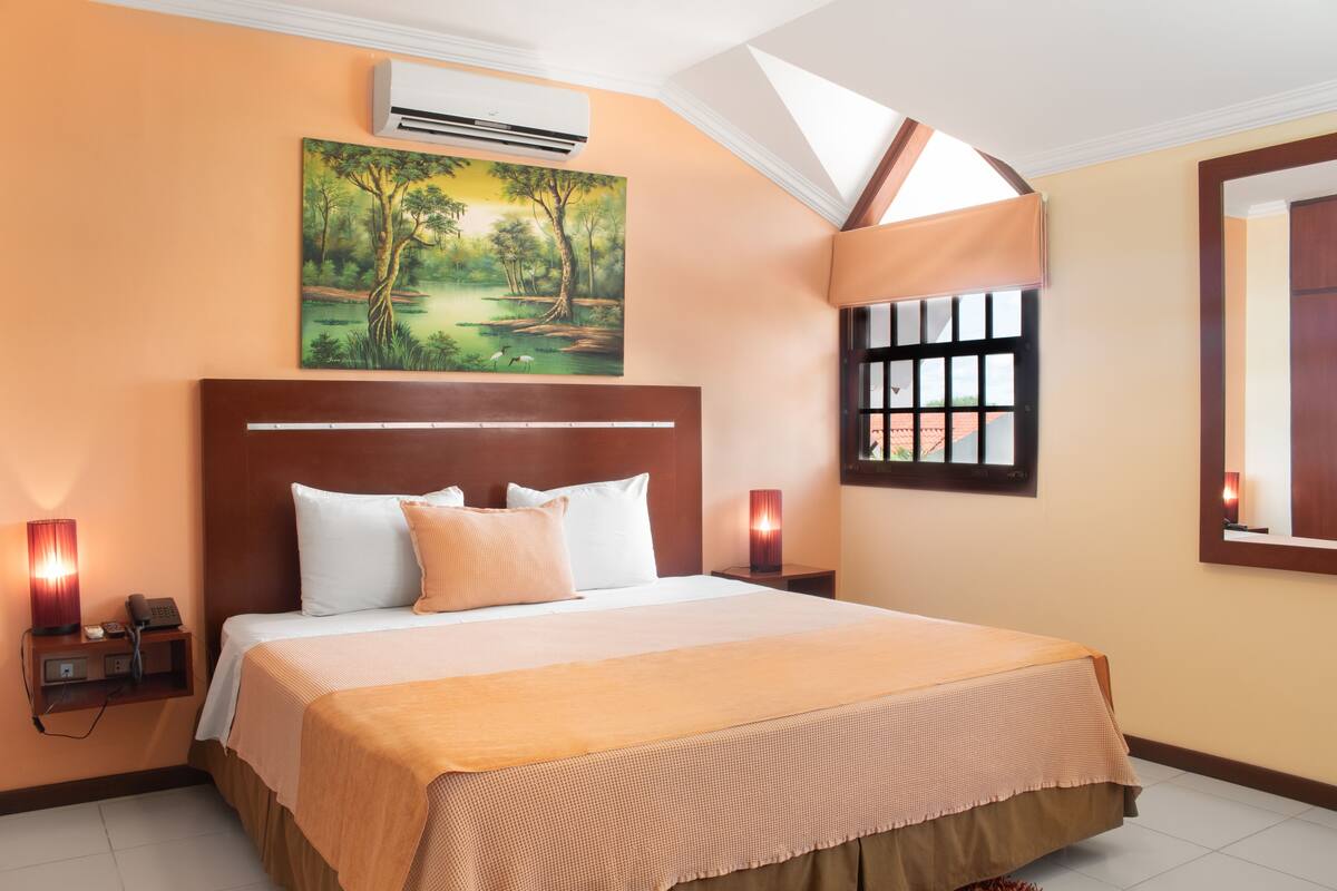 A hotel room with a large bed, a painting of a forest, an air conditioner, a nightstand with a lamp, and a window with a view.