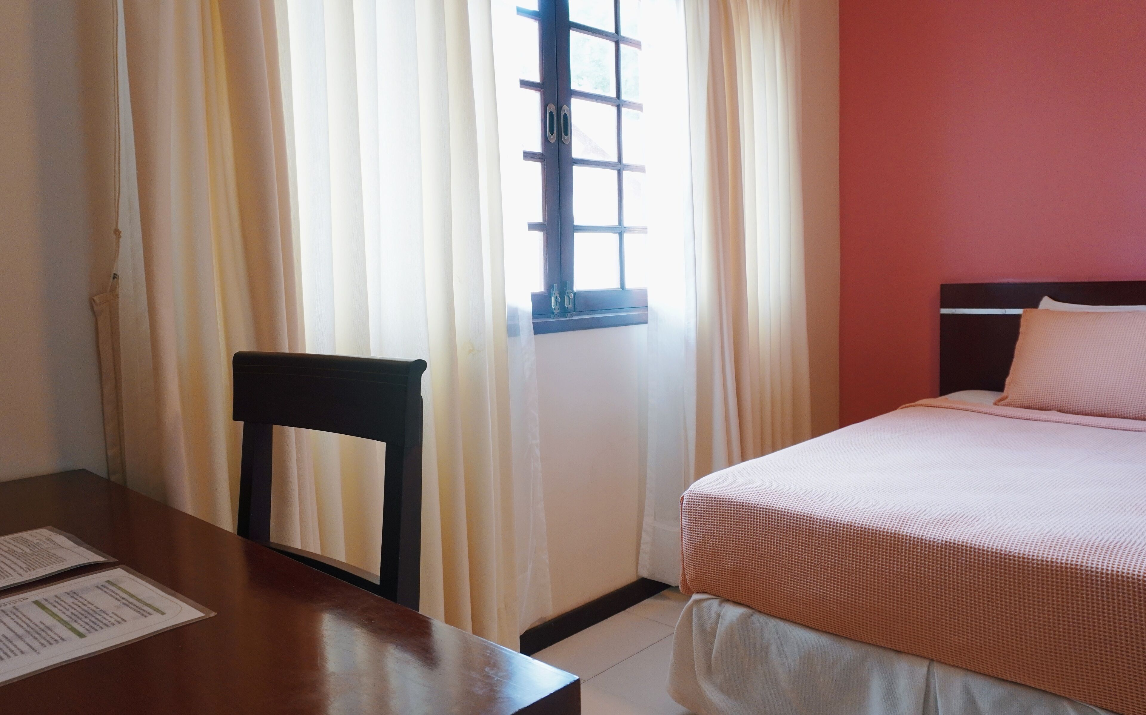 A hotel room with a bed, a wooden chair, a table with a menu, and a window with curtains.