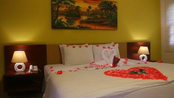 Una habitación de hotel con cama, dos mesitas de noche con lámparas, un cuadro en la pared y pétalos de rosa sobre la cama.