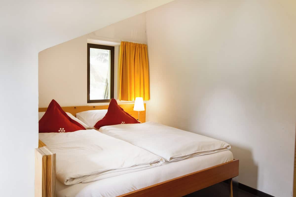 Cama doble con sábanas blancas y almohadas rojas en una habitación con ventana y lámpara.