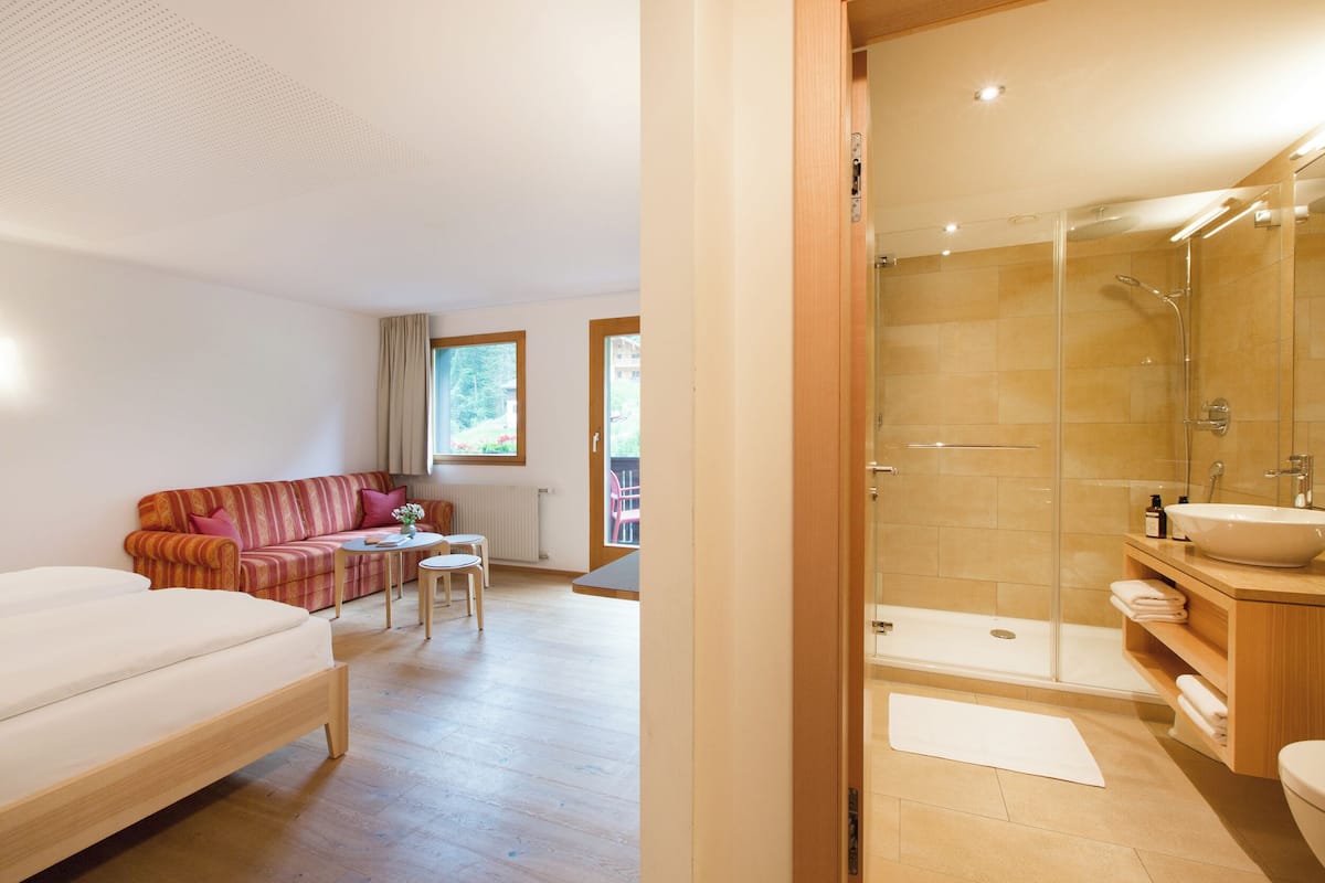 Una habitación de hotel moderna con suelo de madera, un sofá, una mesita y un área de ducha con paredes de vidrio.