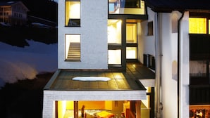 A modern two-story house with a glass facade, a visible interior with a fireplace, and a snowy landscape in the background.