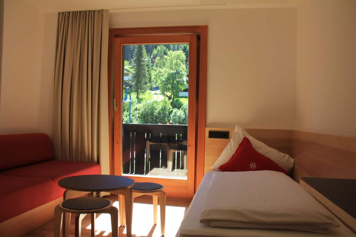 Habitación de hotel con cama, sofá rojo, una mesita y balcón con vista a zonas verdes.