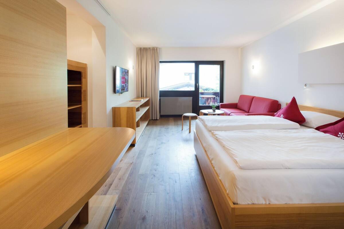 Una habitación de hotel moderna con una cama de madera, un sofá rojo, un escritorio de madera y un ventanal con vistas.