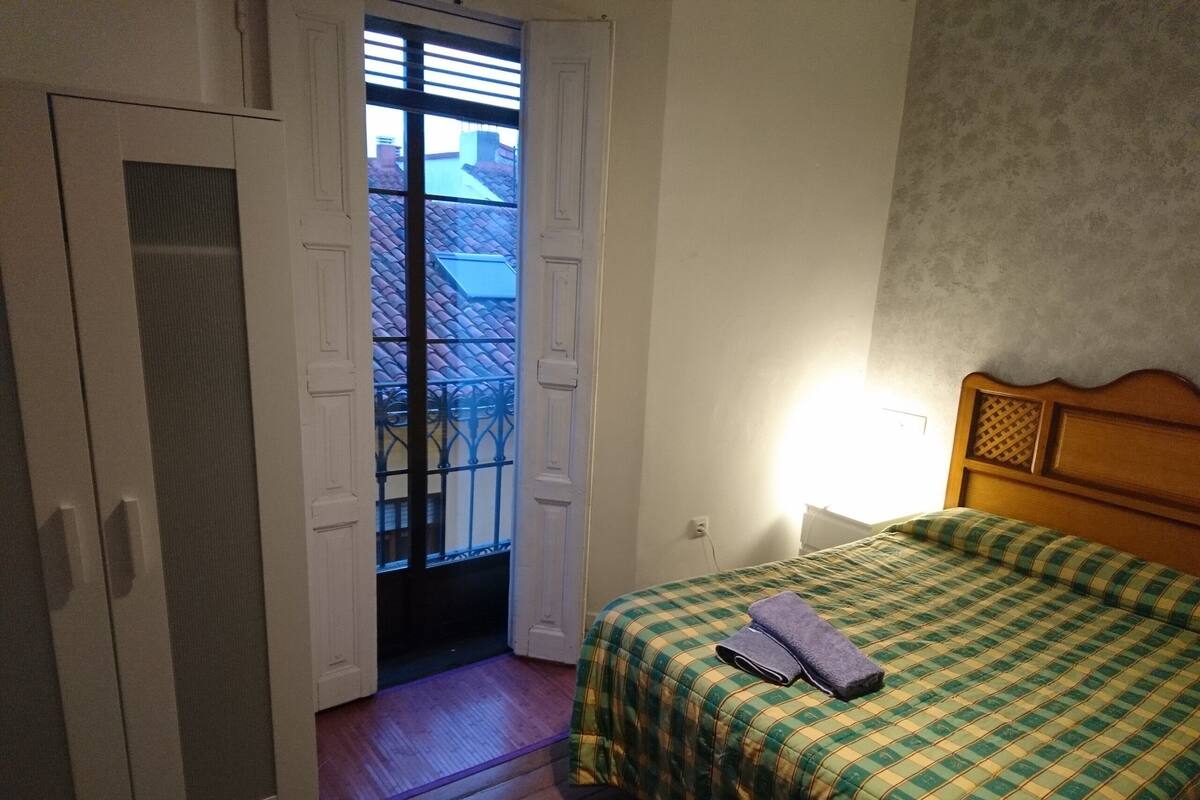 A bedroom with a wooden bed, a green and yellow checkered bedspread, a wooden wardrobe, and a window with a view of rooftops.