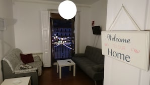 A living room with a sofa, a small table, and a 'Welcome to our Home' sign. A window reveals a cityscape at night. - Apartamentos Plaza Mayor (León)