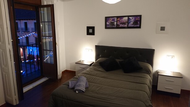 A bedroom with a bed, two bedside tables, a nightstand, a door leading to a balcony, and a wall-mounted picture. - Apartamentos Plaza Mayor (León)