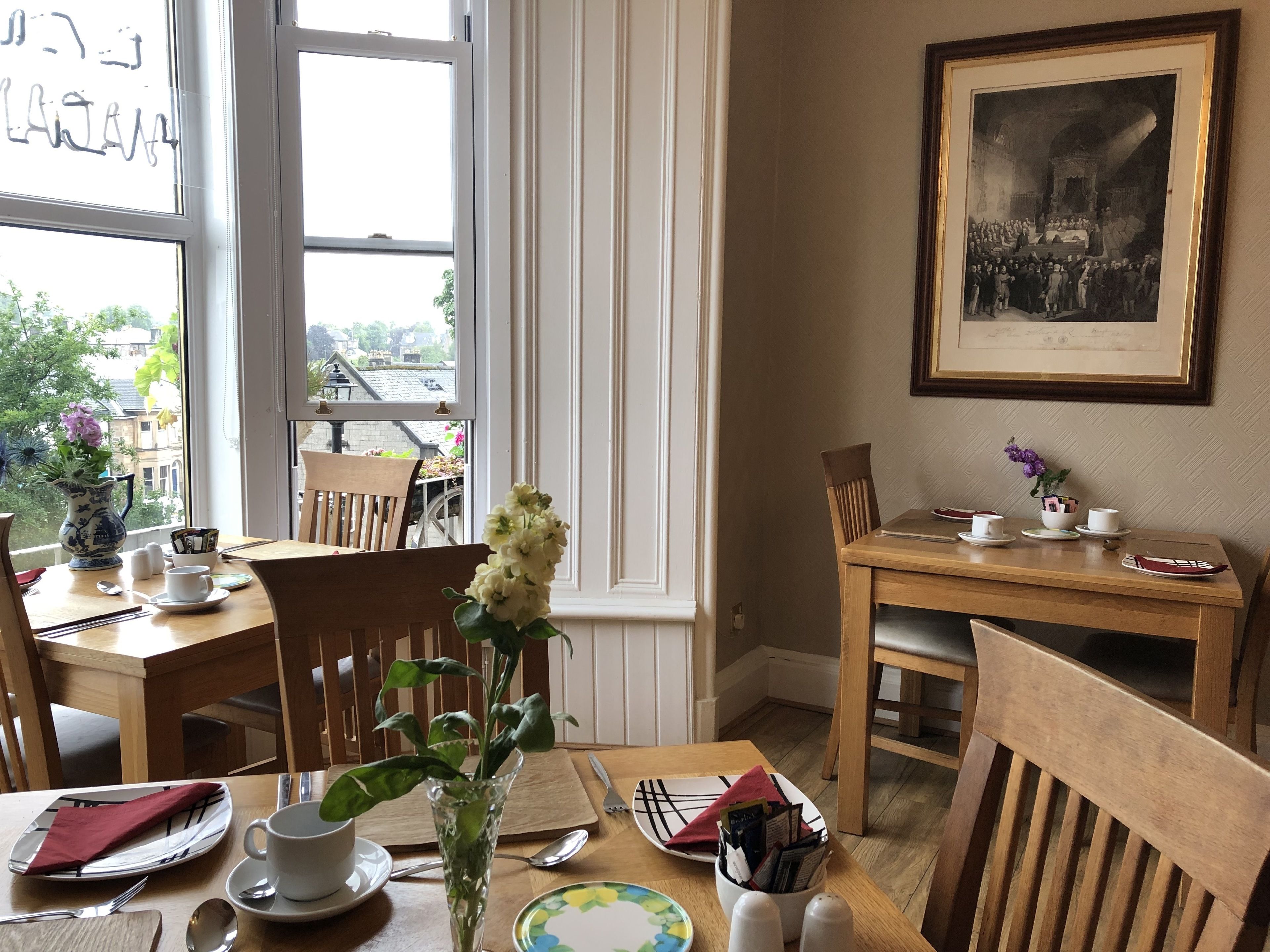 A dining area with a table set for two, a vase with flowers, and a framed historical painting on the wall.