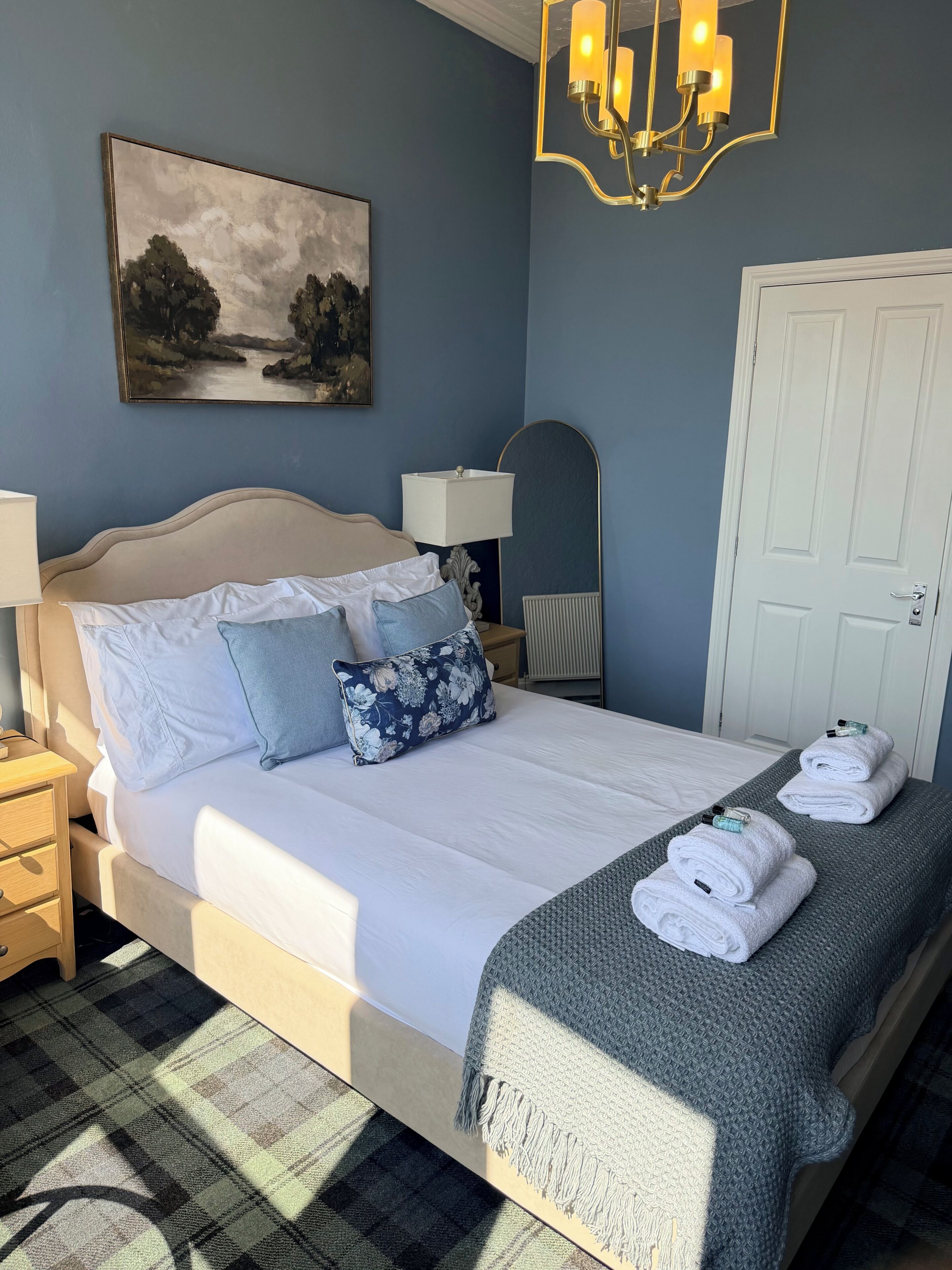 A neatly made bed with pillows, a bedside lamp, a painting on the wall, and a door in the background.