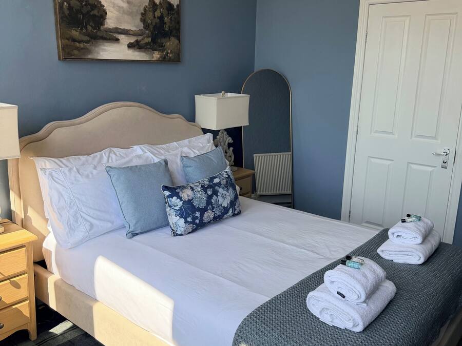 A neatly made bed with pillows, a bedside lamp, a painting on the wall, and a door in the background.