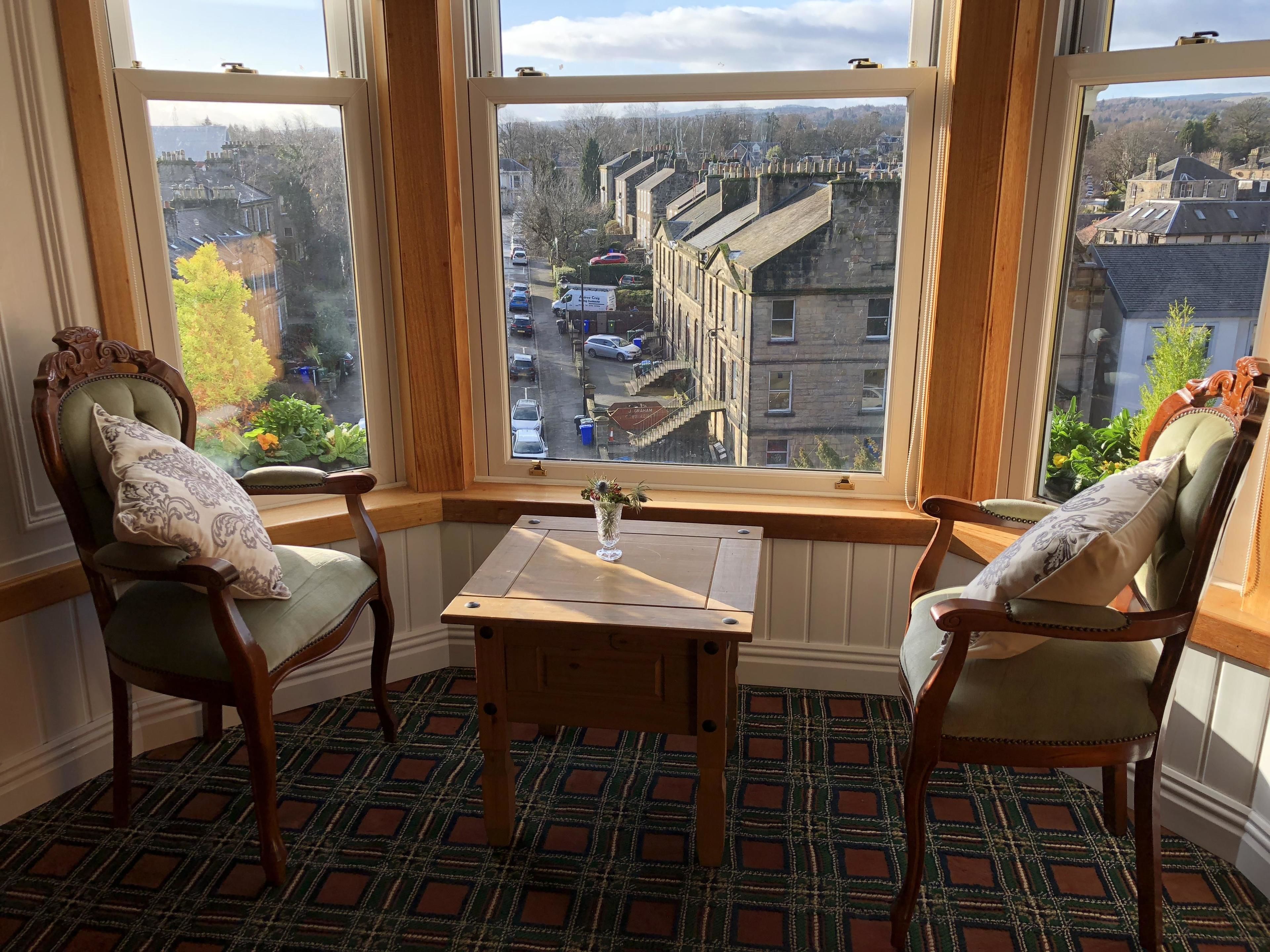 Ein Eckzimmer mit zwei Holzstühlen, einem kleinen Tisch und Blick auf eine Wohnstraße durch große Fenster.