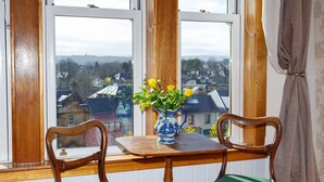 A room with a wooden table and two chairs, a vase with yellow flowers, and a view of houses through a window. - Castle Walk B&B (Stirling)