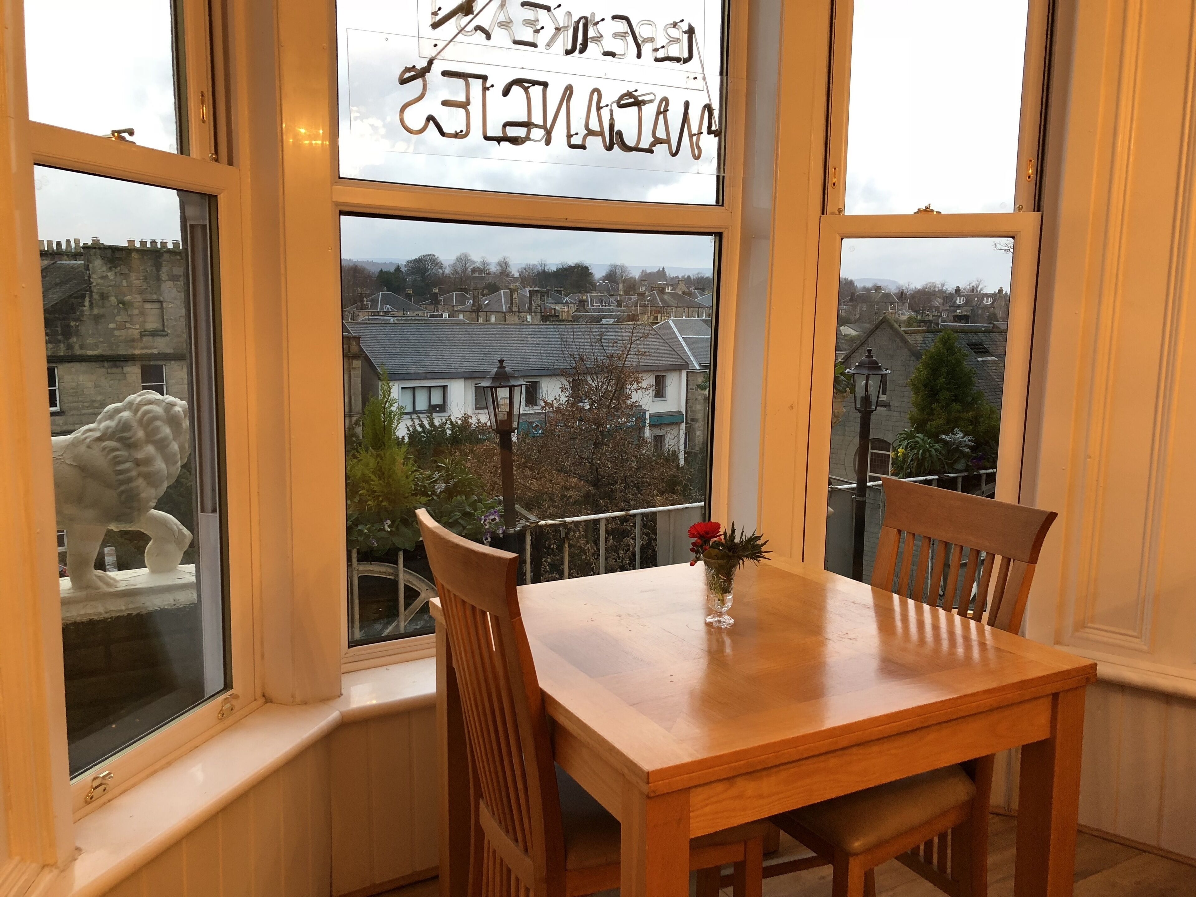 Un coin repas avec une table et des chaises en bois, un vase rempli de fleurs et une vue sur la ville, visible à travers de grandes fenêtres.