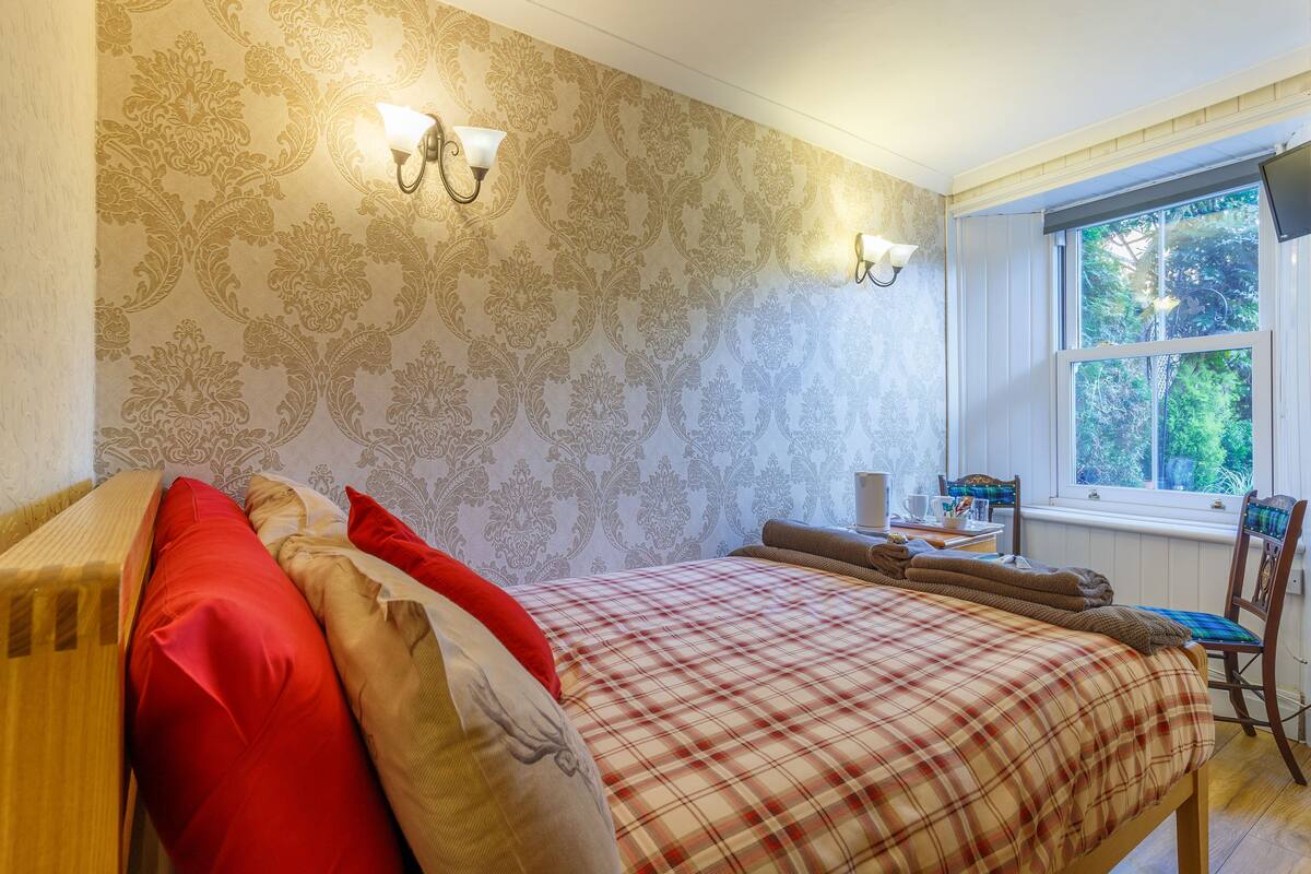 A bedroom with a plaid bedspread, a wooden headboard, and a window with a view of greenery.