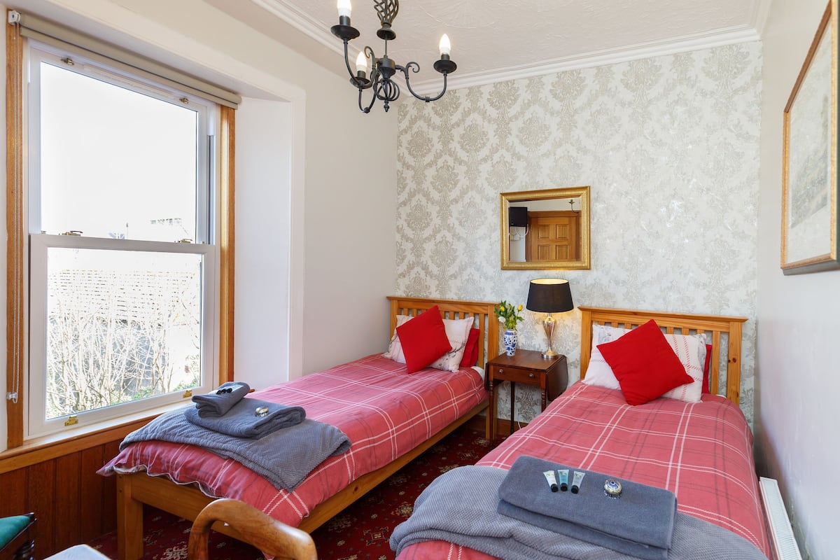 A room with two beds, a window, a chandelier, a mirror, and a small table with a lamp.