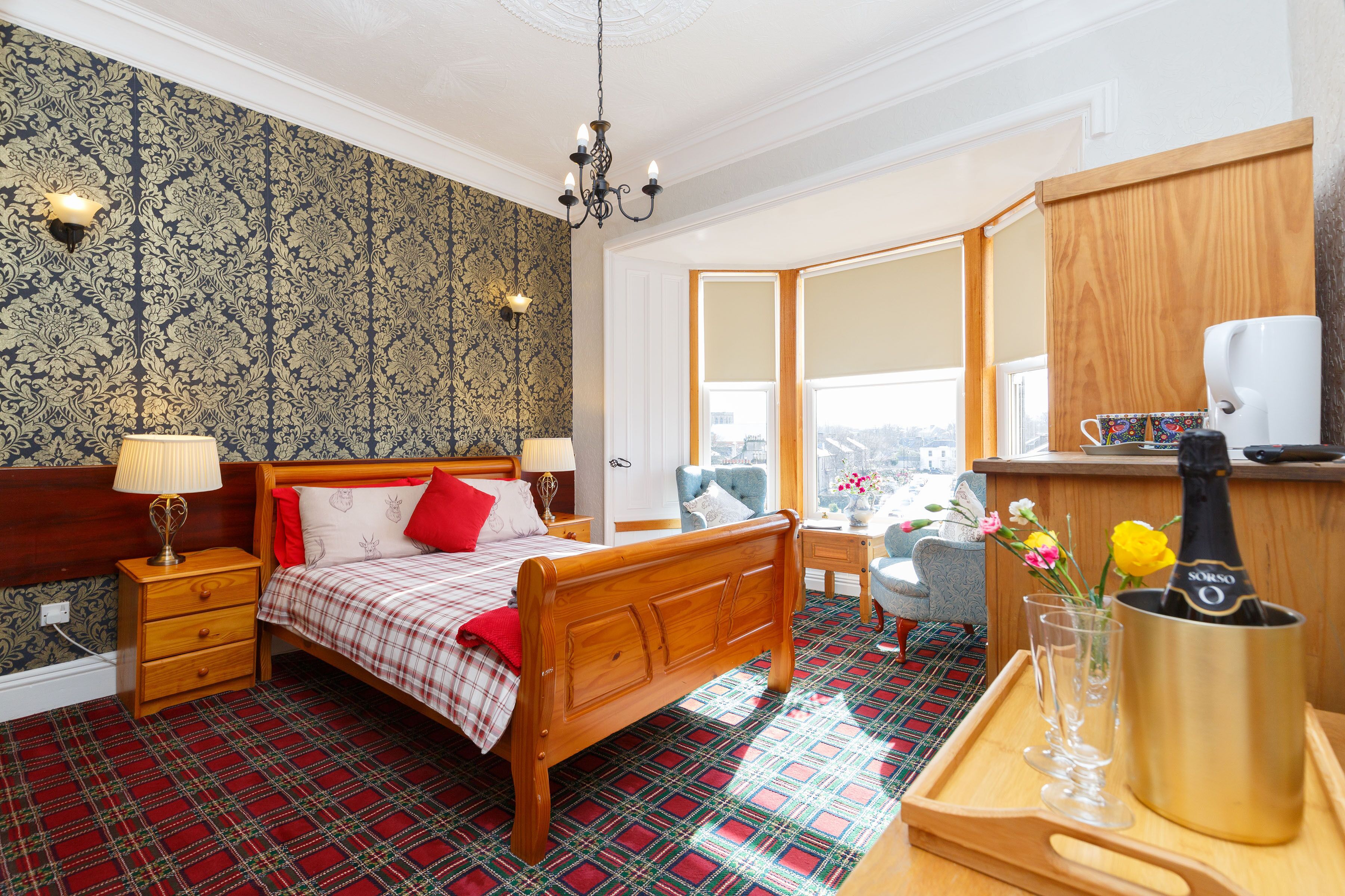 Ein Hotelzimmer mit einem hölzernen Bett, einem Nachttisch mit Lampe, einem kleinen Tisch mit Blumen und einer Minibar, sowie einem großen Fenster mit Blick auf die Stadt.