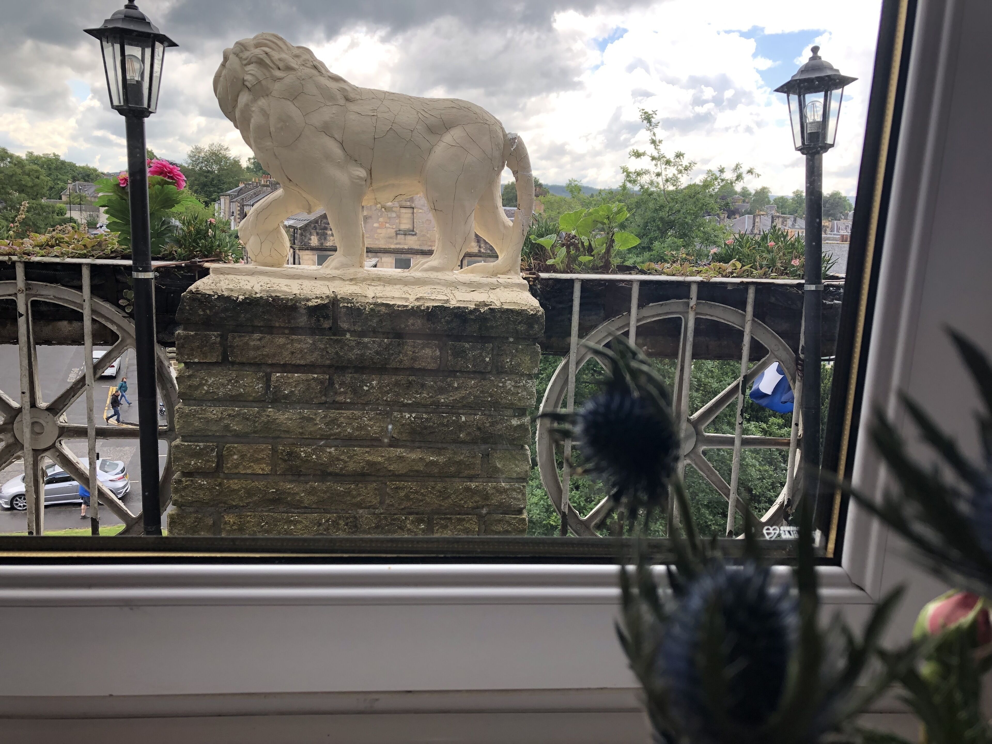 Una estatua de león sobre un pedestal de piedra, vista a través de una ventana con una valla de metal y farolas.