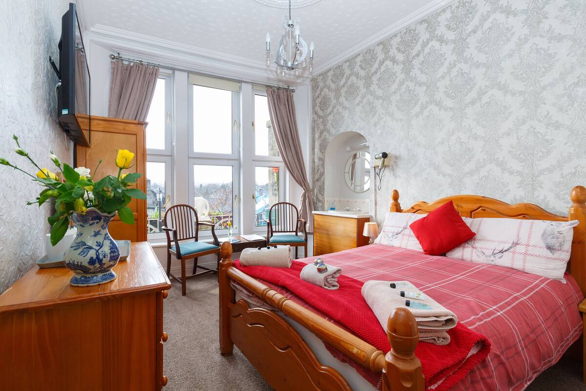 A bedroom with a wooden bed, a vase with flowers, a wooden dresser, a chair, a desk, a mirror, and a window with curtains.