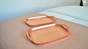 Deux plateaux de couleur cuivrée reposent sur une surface texturée, vraisemblablement un lit ou un canapé, dans une pièce où l’on aperçoit un coussin blanc et un mur uni en arrière-plan.