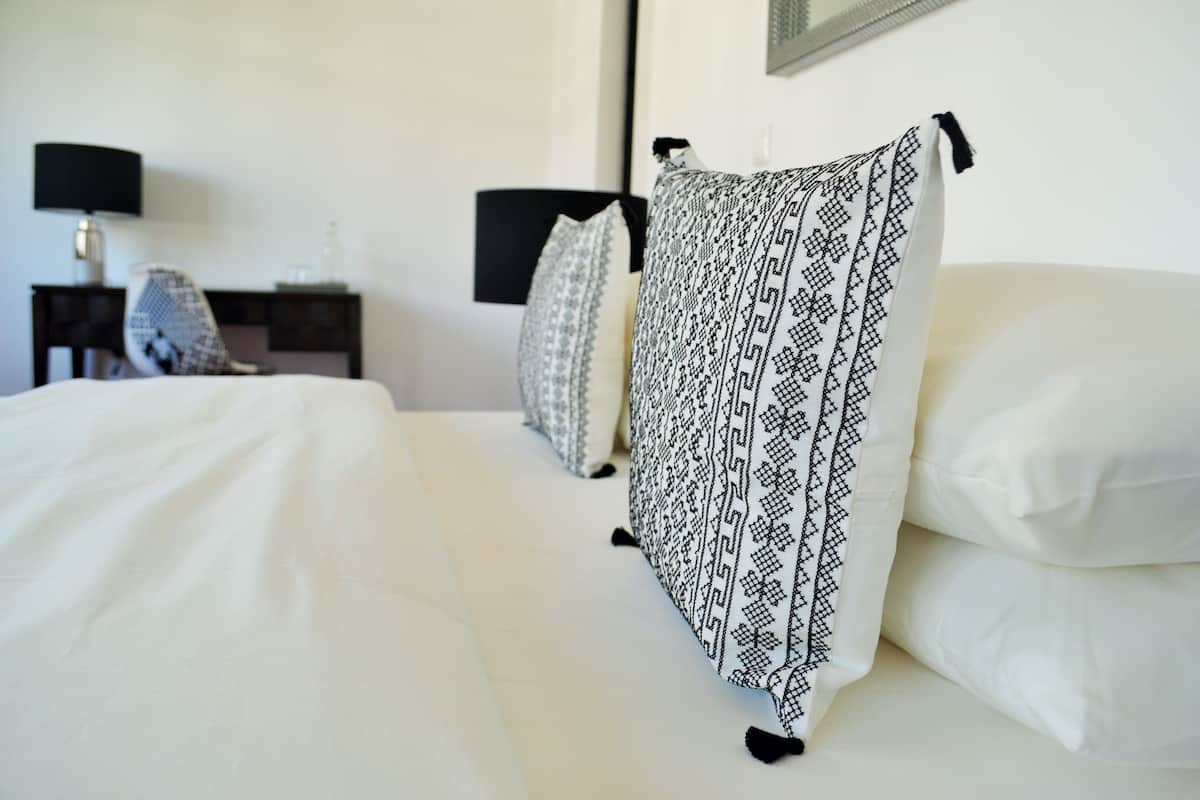 Una cama bien hecha con sábanas blancas y una manta decorativa, una mesita de noche con lámpara y una silla en una habitación de hotel.