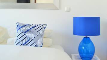 A neatly made bed with a blue bedspread and a patterned pillow, a blue lamp on a white nightstand, and a white wall with a framed picture and a light switch.