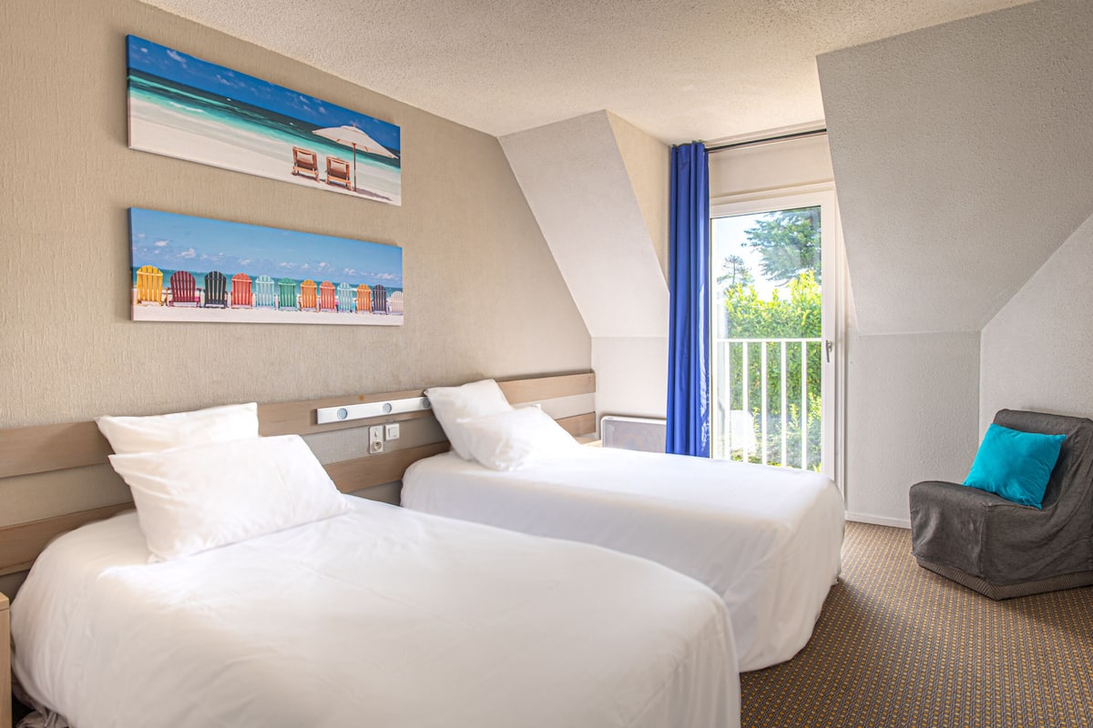 Une chambre d’hôtel avec deux lits, un balcon offrant une vue et un tableau représentant des chaises de plage.