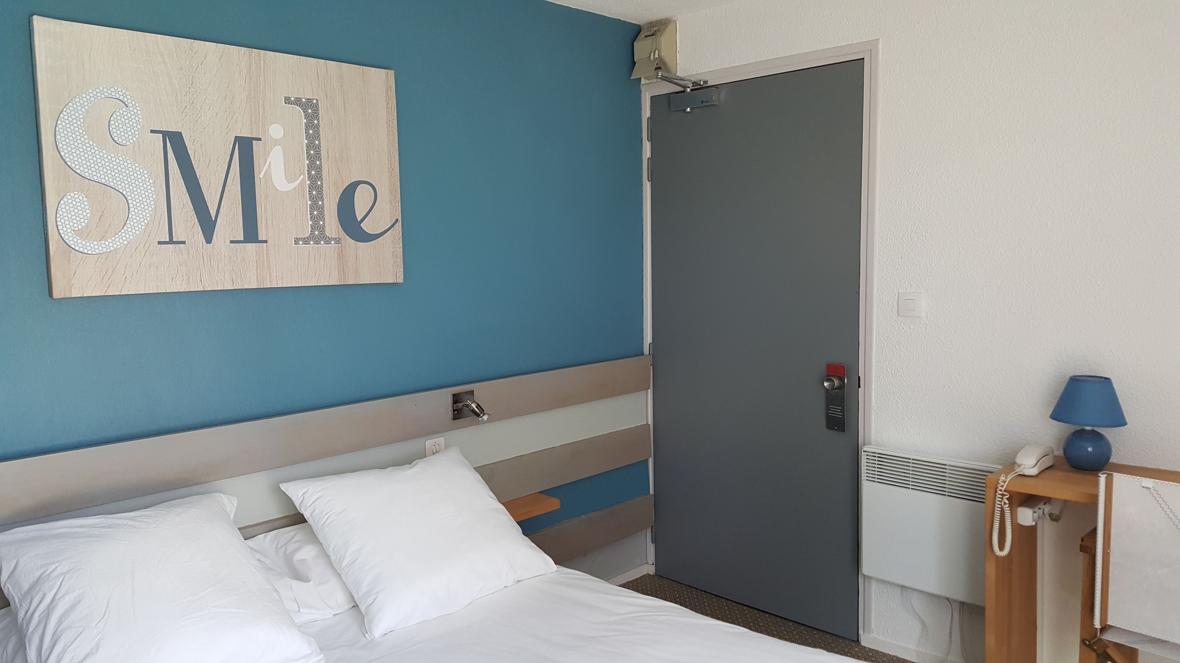 A hotel room with a bed, a door, a bedside table with a lamp, and a wall with a 'Smile' sign.