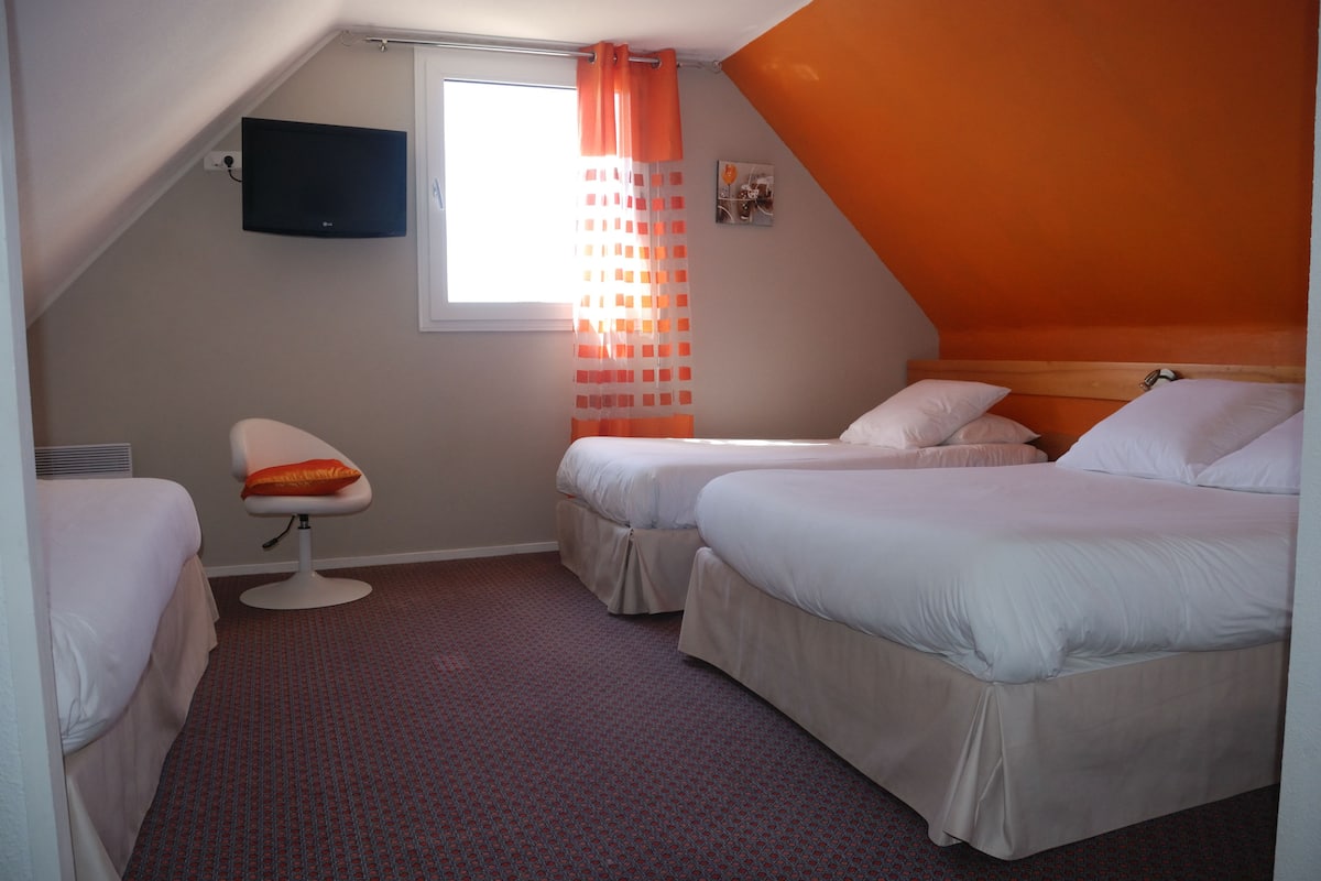 Une chambre d’hôtel avec deux lits, un téléviseur fixé au mur et une fenêtre avec des rideaux orange.